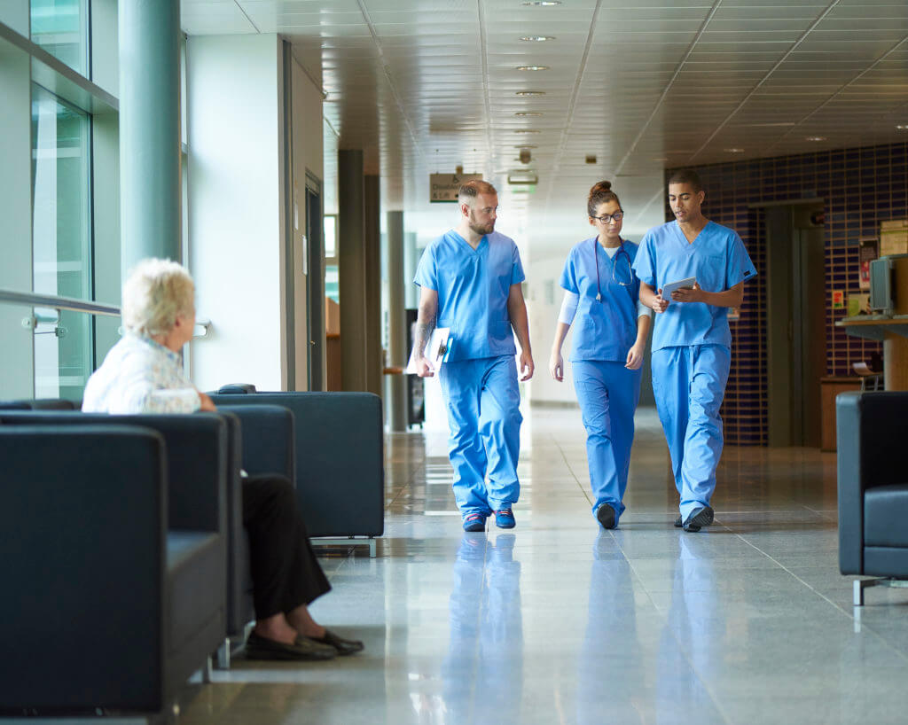 three junior doctors walking along a hospital corridor discussing case and wearing scrubs. A patient or visitor is sitting in the corridor as they walk past .