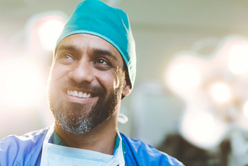 Thoughtful smiling doctor looking away in hospital. Male healthcare worker is wearing scrubs. He is having beard.